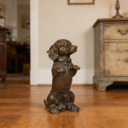 This is a charming bronze sculpture of a dachshund, crafted using the lost-wax casting method. The piece captures the dog in a playful, upright seated pose, with its front paws raised as if begging for attention, conveying a sense of endearing curiosity and eagerness. The rich, warm bronze patina accentuates the dog’s distinctive long body, short legs, and floppy ears, as well as the detailed texture of its short coat and the buckle on its collar.