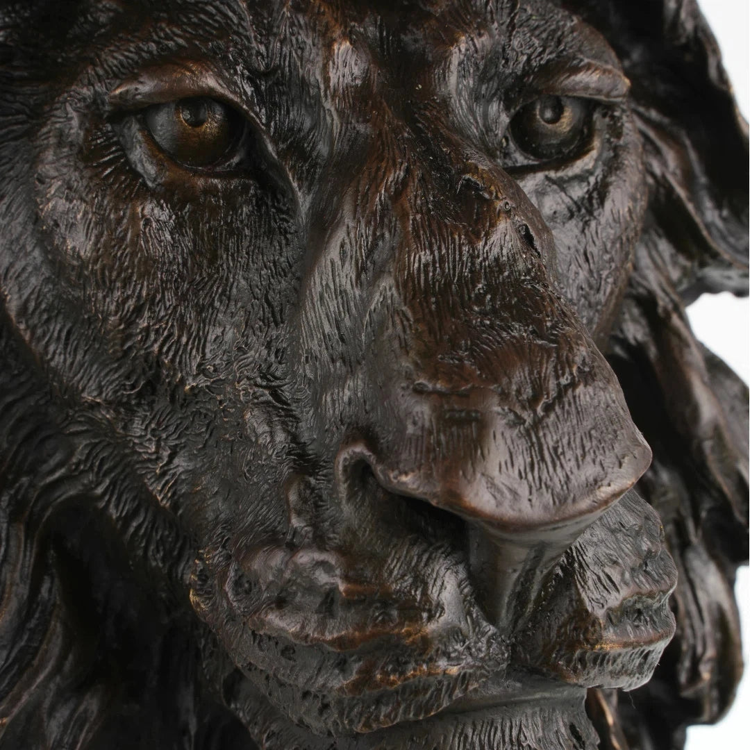 This is a powerful bronze bust of a lion, crafted using the lost-wax casting method. The sculpture focuses on the lion’s head and upper neck, capturing the animal in a moment of intense focus and regal authority. The deep, rich bronze patina accentuates the hyper-realistic details of the lion’s face—from the piercing gaze of its eyes and the rough texture of its whiskers to the intricate, flame-like folds of its mane, which appear to be caught in a powerful gust of wind.